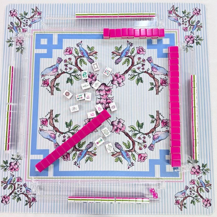 A top-down view of a Mahjong game set on a decorative printed mat. The set includes a blue and white striped background with pink floral and bird motifs, clear acrylic tiles, bright pink racks, and scattered white dice and playing cards.