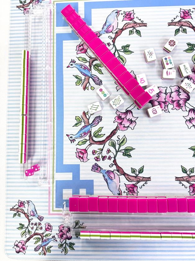 A flat-lay view of a mahjong game set featuring a decorative backboard with pink cherry blossom branches and blue birds, pink tile racks, white tiles with Chinese characters, and two striped pens on a white background.