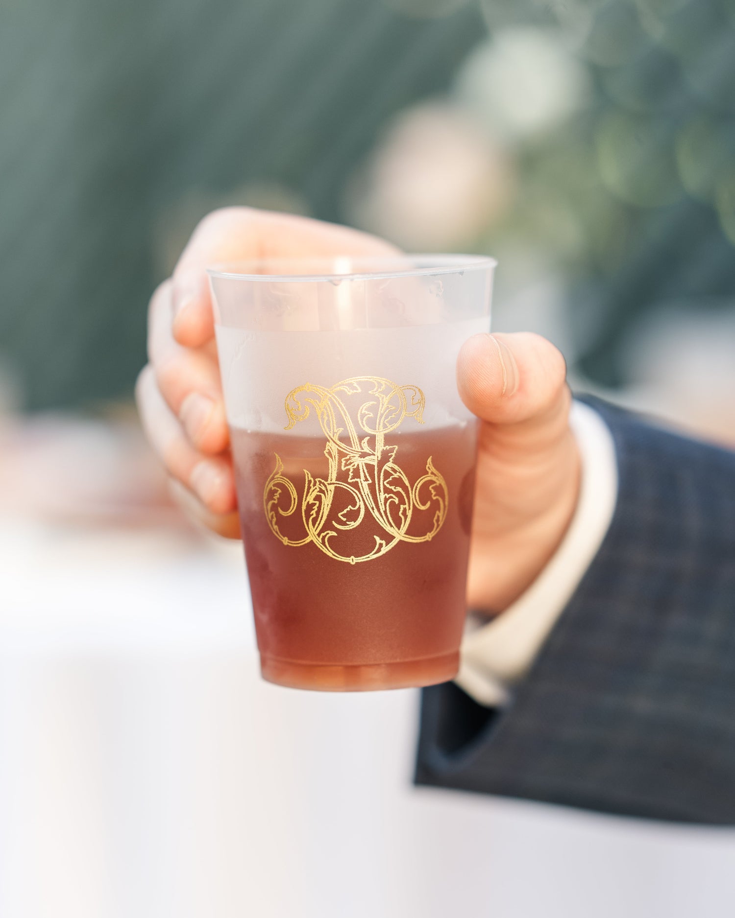 A hand holding a clear plastic cocktail coupe cup filled with a reddish-brown drink, featuring a gold monogram design. The person holding the cup is wearing a dark plaid suit jacket and a white shirt cuff, with a blurred outdoor background.