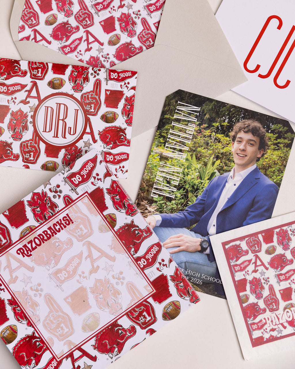 A styled flat-lay of personalized red and white printed stationery and a printed photo card on a light beige background. The stationery includes patterned envelopes, a letterhead, and a memo pad with red foam finger and football motifs, alongside a portrait photo of a young man in a blue blazer.