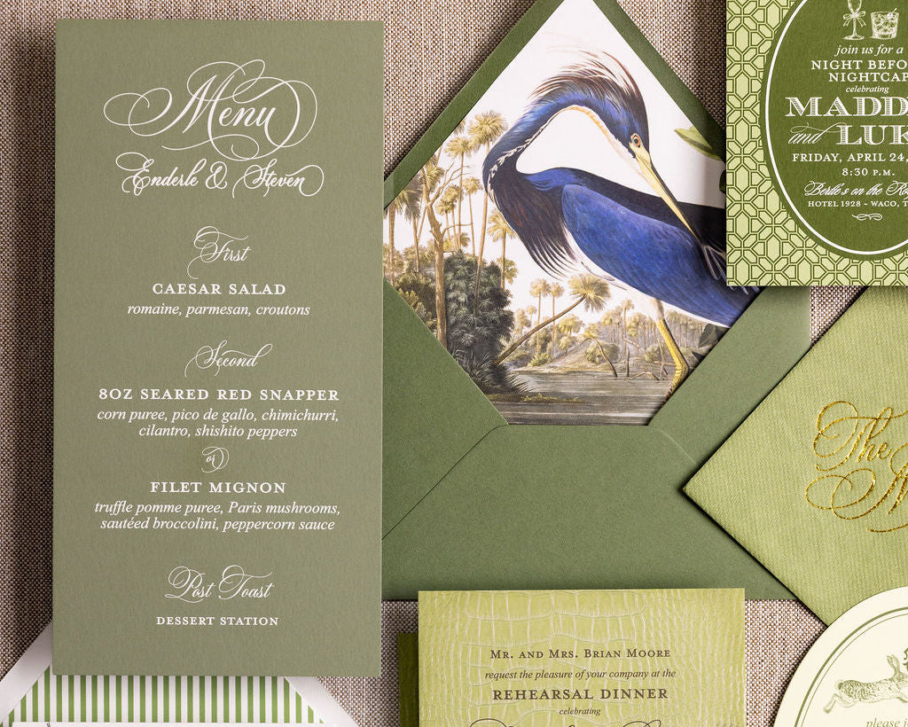 A coordinated stationery set arranged on a textured beige fabric background. The set includes an olive green menu card, a green invitation card with gold script, a green envelope, a bird illustration insert, a white and green round label, and a white illustrated card with guitar and floral artwork.