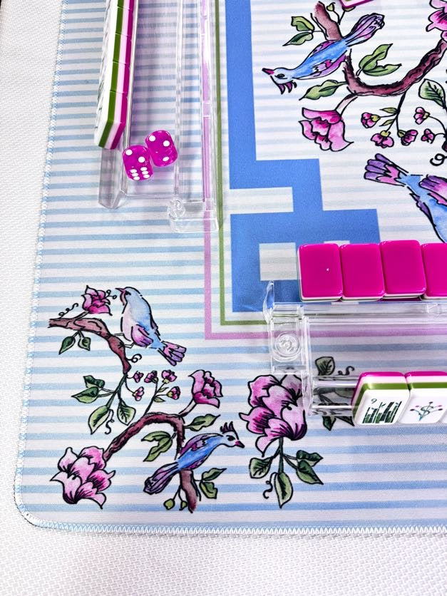 A close-up of a decorative mahjong mat featuring a blue and white striped background with pink flowers and blue birds, alongside matching pink tiles and clear scoring sticks.