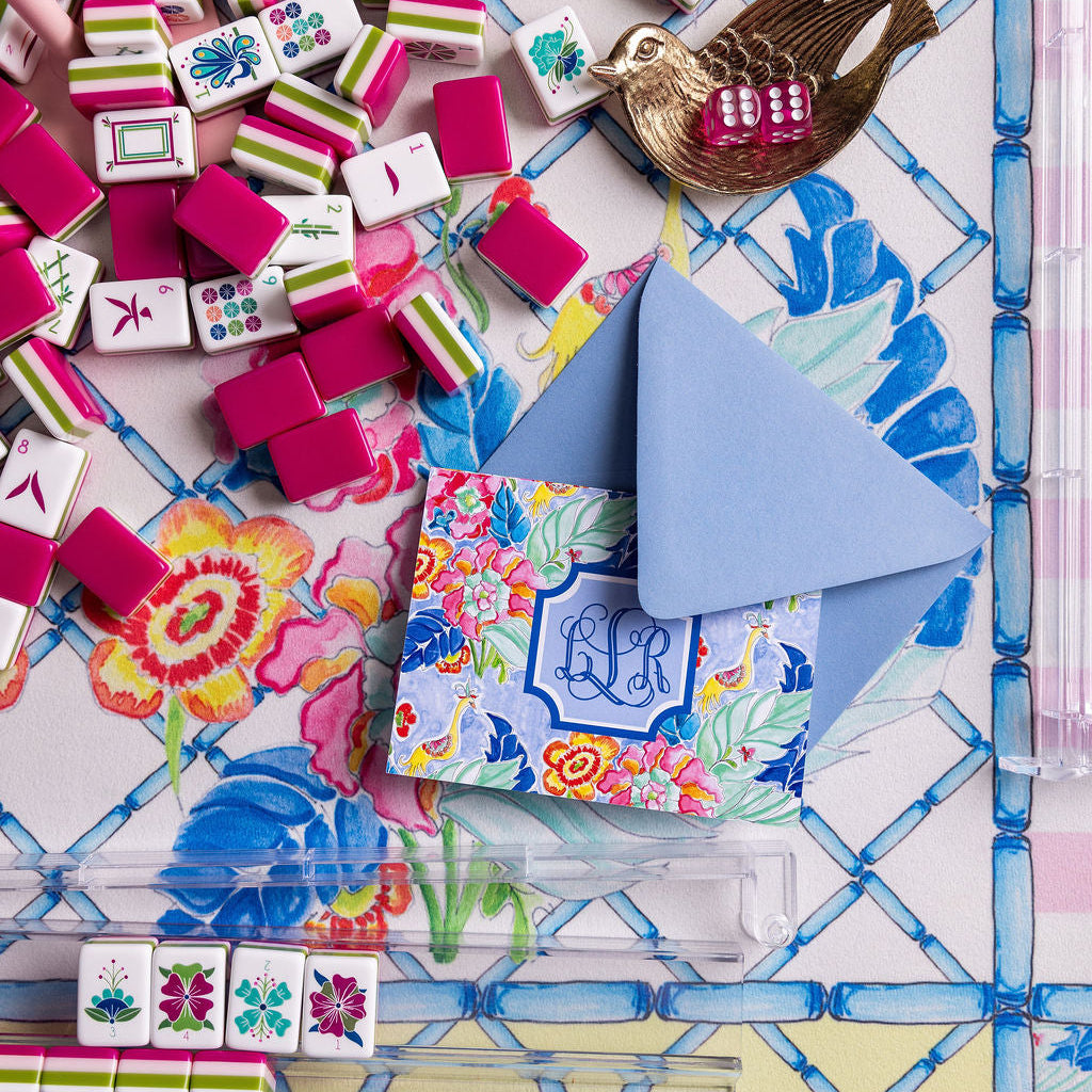 A styled flat-lay of a mahjong set with many pink, green, white, and blue tiles scattered across a floral and geometric patterned mat. A folded blue envelope with a monogram 'LH' sits among the tiles, along with a small gold bird-shaped accessory and a clear plastic rack holding a few tiles.