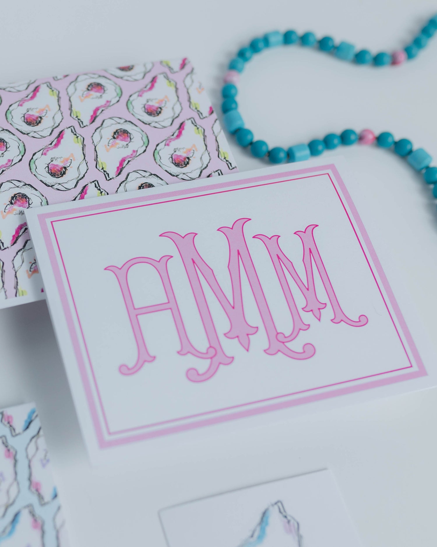 A styled flat-lay of patterned stationery and accessories on a white surface, including a central card with pink monogram letters 'MMM' in a pink border, another card with an oyster shell pattern, a beaded turquoise necklace, and a small oyster shell dish.