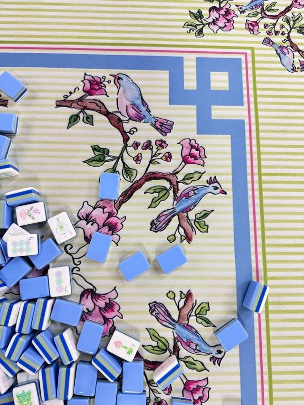 A close-up of a decorative mahjong tile layout featuring blue and white tiles scattered over a printed mat with birds, flowers, and striped borders.
