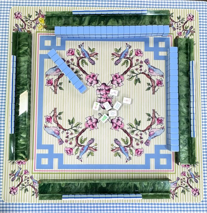 A square decorative game board or card set photographed from above, featuring a floral and bird pattern with blue tiles arranged around the edges and white dice in the center on a blue-and-white checkered surface.