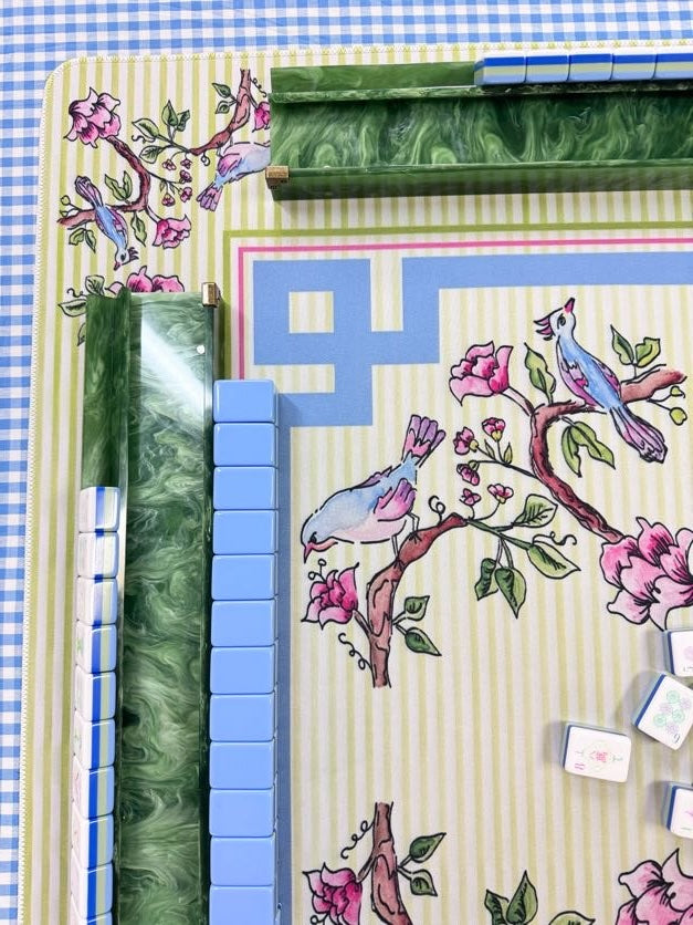 A close-up of a decorative mahjong set with a cream striped mat printed with pink flowers and blue birds, green marbled tile holders, blue-backed tiles, and a blue-and-white gingham surface underneath.