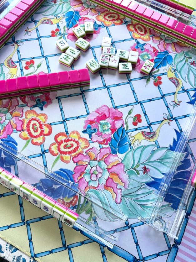 A close-up of a Mahjong game set with a colorful floral and bird-patterned board, pink-backed tiles, scattered white tiles with green and red markings, and a clear acrylic rack.
